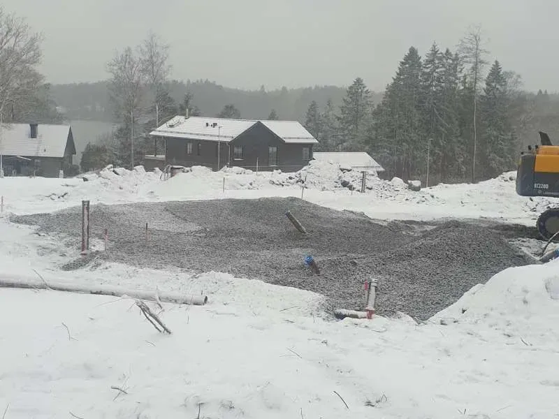 Snötäckt tomt med grusbädd och en grävmaskin i kanten